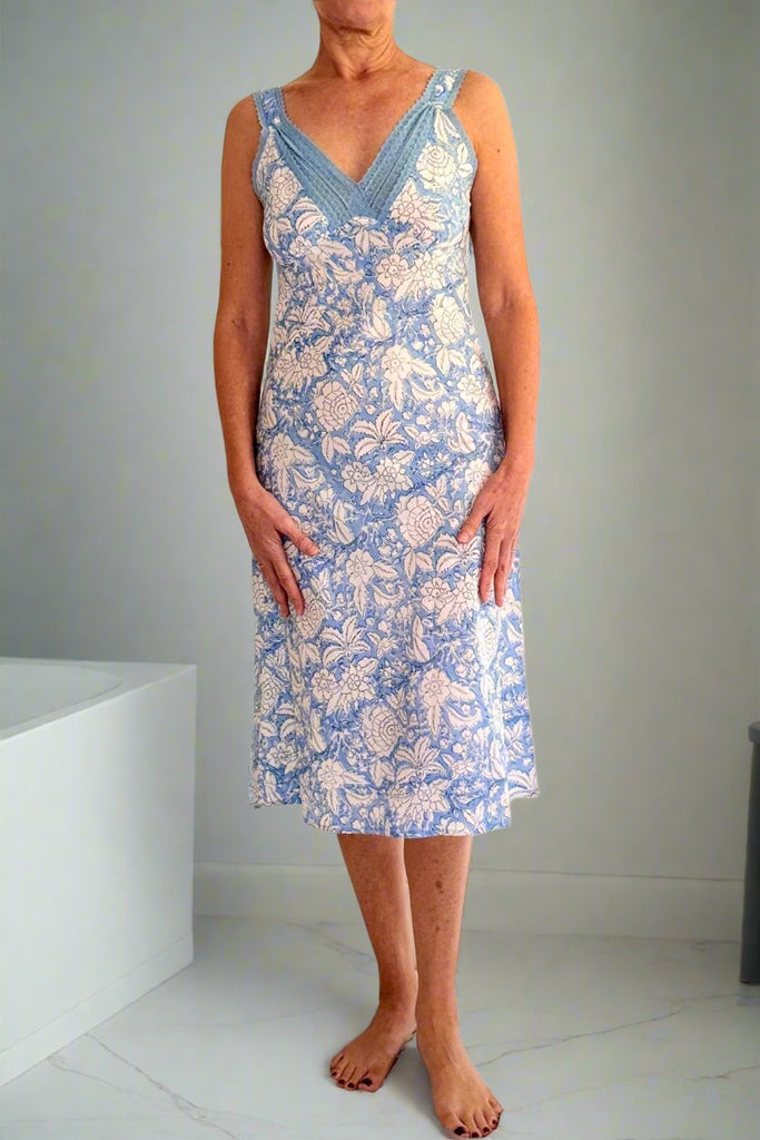 Woman wearing blue and white floral print cotton strappy, fitted nightdress with blue lace trim standing next to a bath